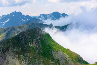 Yüksek dağlarda çarpıcı bir manzara. Yamaçlarda çimenler ve kar lekeleri. Uzakta kayalık bir tepe. Vadiden yükselen bulutlar