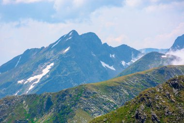 Yüksek dağlarda çarpıcı bir manzara. Yamaçlarda çimenler ve kar lekeleri. Uzakta kayalık bir tepe. Yüksek nem ile bulutlu bir gün