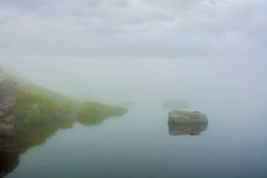 Gölde sis var. Taşlı çimenli bir sahil. Bulutlu gökyüzü. gizemli doğa. kötü görünürlük kavramı