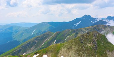Yüksek dağlarda çarpıcı bir manzara. Yamaçlarda çimenler ve kar lekeleri. Uzakta kayalık bir tepe. Yüksek nem ile bulutlu bir gün