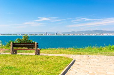 Güneşli sahil kıyısında bir bank. Sahilde asfaltlı patikası olan boş bir parktan güzel bir gökyüzü manzarası. Şehir ve dağ, bulutlu mavi gökyüzünün altında.