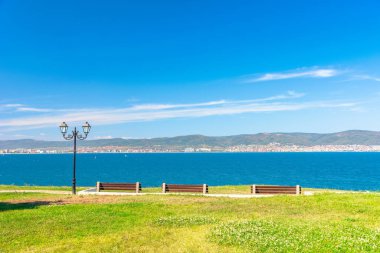 Güneşli sahil kıyısında üç bank. Sahilde asfaltlı patikası olan boş bir parktan güzel bir gökyüzü manzarası. Şehir ve dağ, bulutlu mavi gökyüzünün altında.
