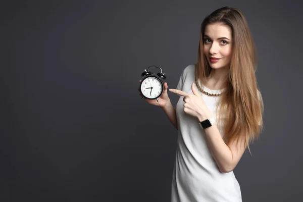 Mujer delgada joven que apunta al reloj de alarma sobre fondo gris — Foto de Stock