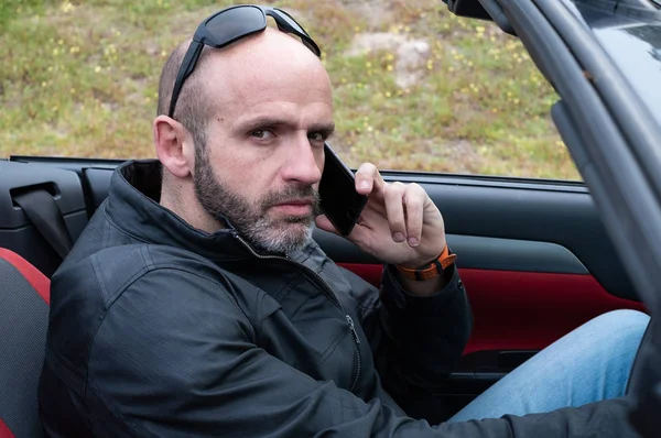 Middle-age bald and serious man with sunglasses and leather jacket talking on his smart phone in his convertible car
