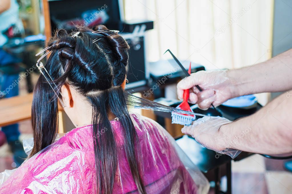 The hands of the hairdresser apply color to dark long hair