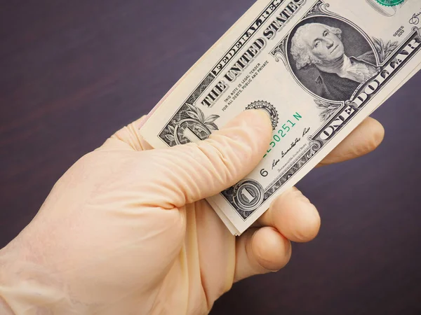 Et rengi lastik eldivendeki bir el bir tomar Amerikan parası tutar. Bir dolarlık banknotun üstüne. Koyu kahverengi arka plan. Koronavirüse karşı önlem, önlem, salgın sırasında COVID-2019