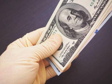 Renksiz tıbbi lateks eldivenli bir el Amerika 'nın parasını karşılar. Yukarıda bir dolarlık banknot var. Koyu kahverengi arka plan. Koronavirüse karşı önlem, önlem, salgın sırasında COVID-2019