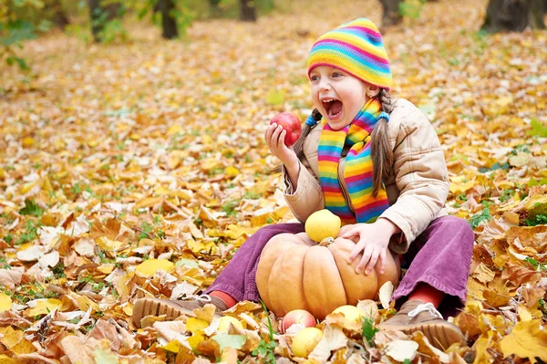 Menina criança na floresta de outono, sente-se em folhas amarelas, coma maçã e abóbora — Fotografia de Stock
