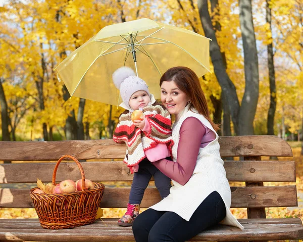 Женщина с ребенком девочка в осеннем городском парке сидеть на скамейке с корзиной яблок и зонтиком и весело, счастливая семья — стоковое фото