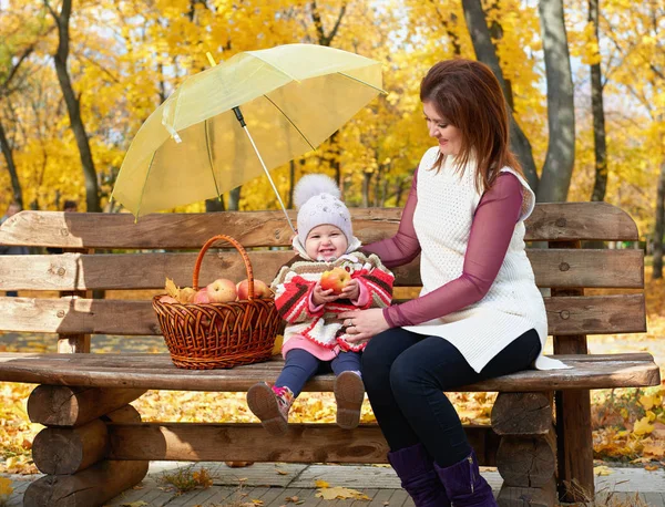Femme avec enfant fille dans le parc de la ville d'automne assis sur un banc avec panier de pommes et parapluie et avoir du plaisir, famille heureuse — Photo