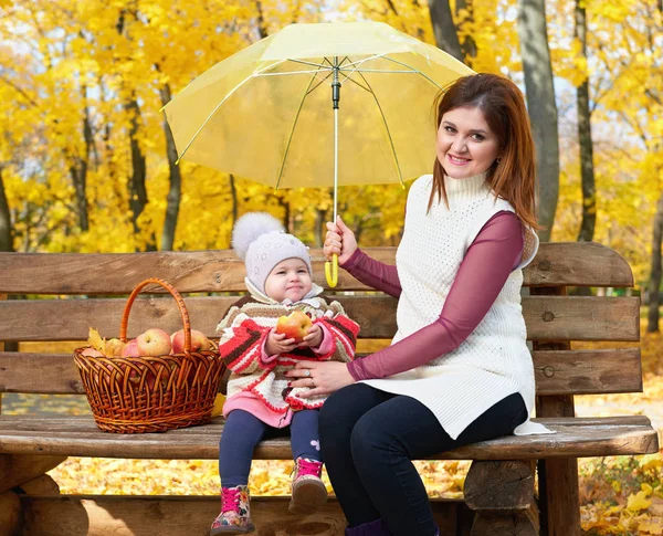 Woman with child girl in autumn city park sit on bench with apples basket and umbrella and having fun, happy family — Stock Photo, Image
