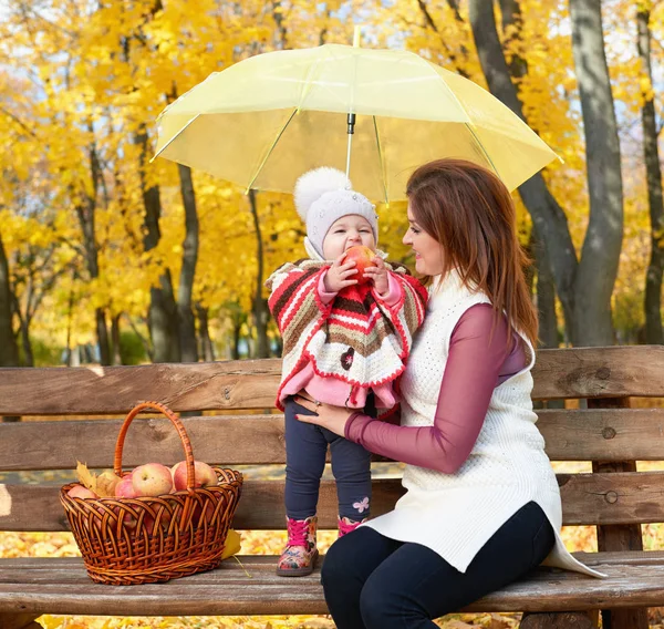 Mujer con niña en el parque de otoño de la ciudad sentarse en el banco con la cesta de manzanas y paraguas y divertirse, familia feliz —  Fotos de Stock