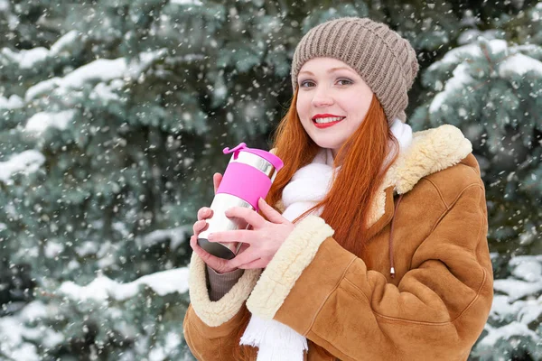 Hermosa mujer bebiendo una bebida caliente y mantener caliente en invierno al aire libre, abetos nevados en el bosque, pelo largo y rojo, con un abrigo de piel de oveja — Foto de Stock