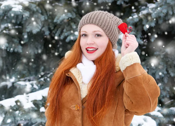 Hermosa mujer en invierno posando al aire libre con juguetes en forma de corazón, concepto de vacaciones, abetos nevados en el bosque, pelo largo y rojo, con un abrigo de piel de oveja — Foto de Stock