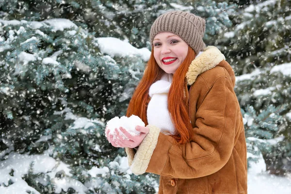 beautiful woman play with snow on winter outdoor, snowy fir trees in forest, long red hair, wearing a sheepskin coat