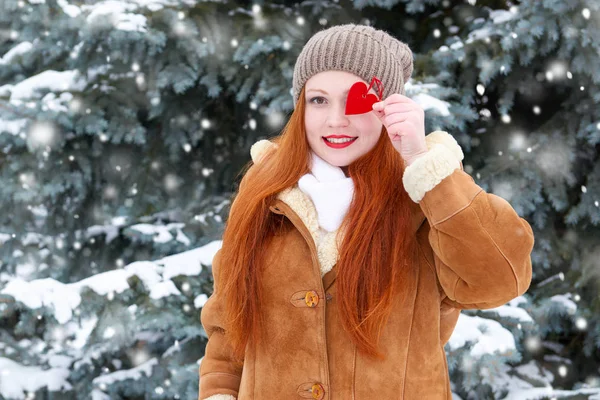 Hermosa mujer en invierno posando al aire libre con juguetes en forma de corazón, concepto de vacaciones, abetos nevados en el bosque, pelo largo y rojo, con un abrigo de piel de oveja — Foto de Stock