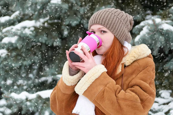 美丽的女人喝喝些热饮料和保暖冬季户外、 雪杉树在森林里，长长的红色头发，穿着一件羊皮大衣 — 图库照片