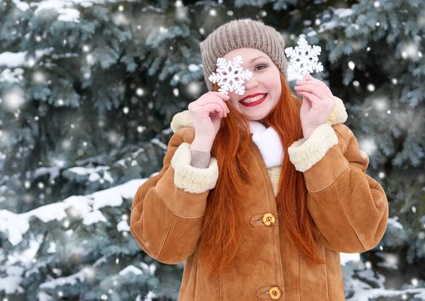 Красивая женщина на зимнем открытом воздухе позирует с большими снежинками игрушки, праздничная концепция, снежные елки в лесу, длинные рыжие волосы, носить пальто овчины — стоковое фото