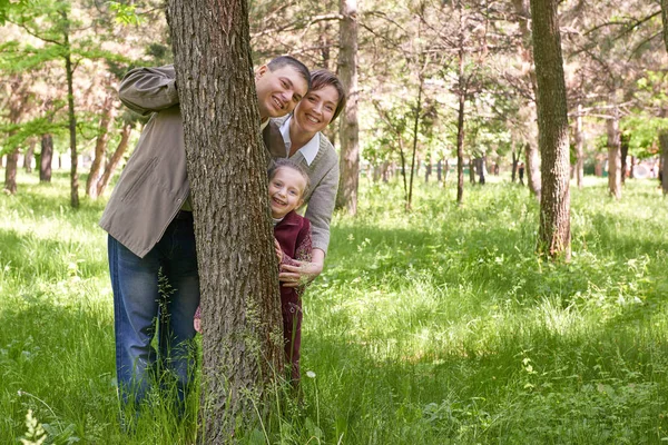 Glückliche Familie und Kind im Sommerpark. Menschen, die sich hinter einem Baum verstecken und spielen. schöne Landschaft mit Bäumen und grünem Gras — Stockfoto