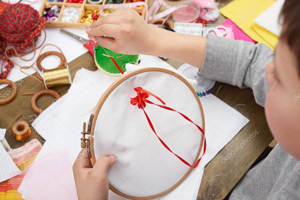 boy embroidered on the hoop, hand closeup and red ribbon on white textile, learns to sew, job training, handmade and handicraft concept