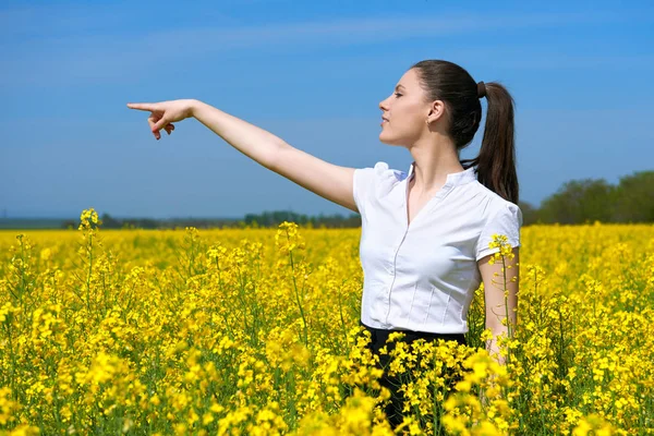 Geschäftsfrau blickt in die Ferne und zeigt auf den Punkt. junges Mädchen im gelben Blumenfeld. schöne Frühlingslandschaft, strahlend sonniger Tag, Raps — Stockfoto