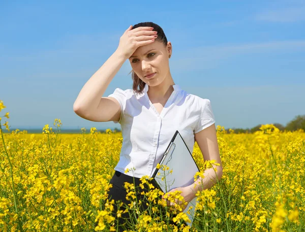 Sad business woman in flower field outdoor with clipboard touch head. Young girl in yellow rapeseed field. Beautiful spring landscape, bright sunny day — Stock Photo, Image