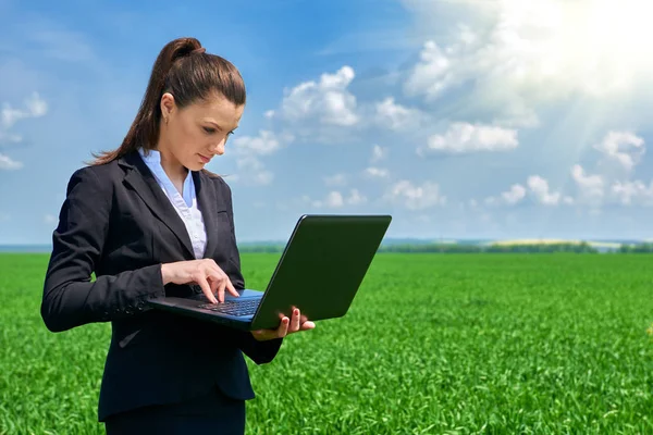 Mulher de negócios no campo de grama verde trabalho ao ar livre no laptop. Menina vestida de terno preto. Bela paisagem de primavera com céu nublado, dia ensolarado brilhante — Fotografia de Stock