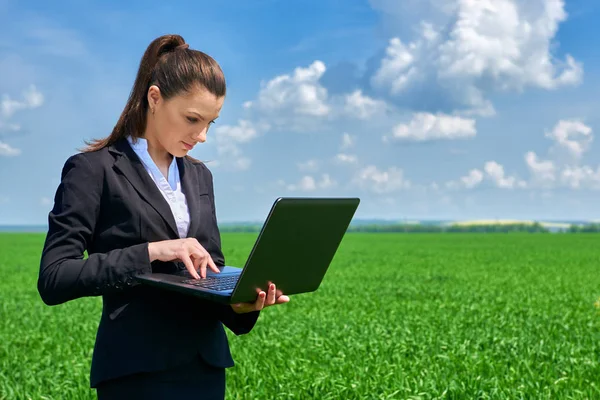 Mujer de negocios en el campo de hierba verde trabajo al aire libre en el ordenador portátil. Chica joven vestida con traje negro. Hermoso paisaje de primavera con cielo nublado, día soleado brillante — Foto de Stock