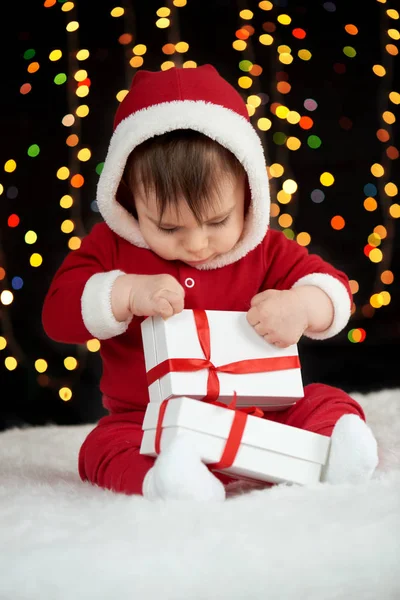 Bebé desempaquetar cajas de regalo con decoración de Navidad, vestido como Santa Claus, luces de arco sobre fondo oscuro, concepto de vacaciones de invierno — Foto de Stock
