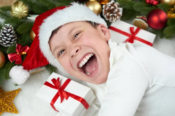Хлопчик, розважається з різдвяним прикрасою, виразом обличчя та щасливими емоціями, одягнений у капелюх Санти, лежить на білому фоні хутра, концепція зимових свят — стокове фото