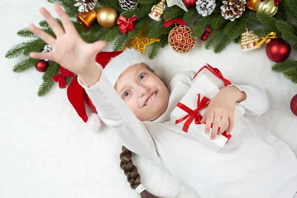 Child girl having fun with christmas decoration, face expression and happy emotions, dressed in santa hat, lie on white fur background, winter holiday concept — Stock Photo, Image