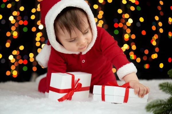 Baby unpack gift boxes with christmas decoration, dressed as Santa, boke lights on dark background, winter holiday concept — Stock Photo, Image