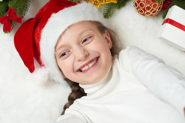 Menina se divertindo com decoração de Natal, expressão facial e emoções felizes, vestida com chapéu de Papai Noel, mentira no fundo de pele branca, conceito de férias de inverno — Fotografia de Stock