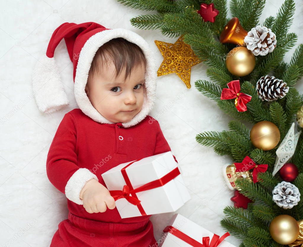 baby portrait in christmas decoration, dressed as Santa, lie on fur near fir tree and play with gifts, winter holiday concept