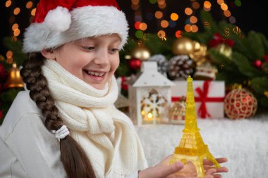 çocuk kız portre Eyfel Kulesi heykelcik ve Noel dekorasyonu ile ışıklar, yüz ifadesi ve mutlu duygular, koyu arka plan giymiş Noel Baba şapkası, kış tatil kavramı