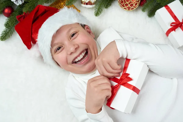 男の子の子供のクリスマスの装飾、表情と幸せな感情で楽しんで、サンタの帽子に身を包んだ、白い毛皮の背景、冬の休日の概念にうそをつく — ストック写真