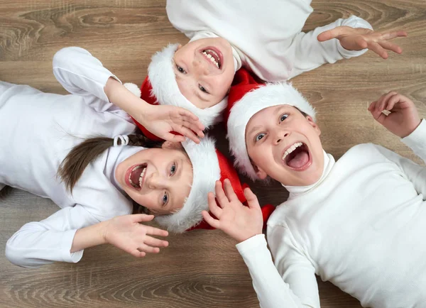 Tres niños en los sombreros de Santa que mienten en el fondo de madera, teniendo emociones divertidas y felices, concepto de vacaciones de invierno — Foto de Stock