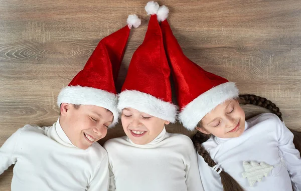 Tres niños en los sombreros de Santa que mienten en el fondo de madera, teniendo emociones divertidas y felices, concepto de vacaciones de invierno —  Fotos de Stock