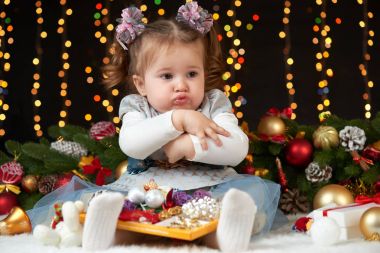 child girl hides her toys, portrait in christmas decoration, winter holiday concept, dark background with illumination and boke lights clipart