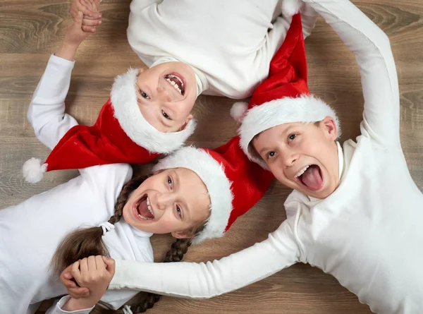 Tres niños en los sombreros de Santa que mienten en el fondo de madera, teniendo emociones divertidas y felices, concepto de vacaciones de invierno — Foto de Stock