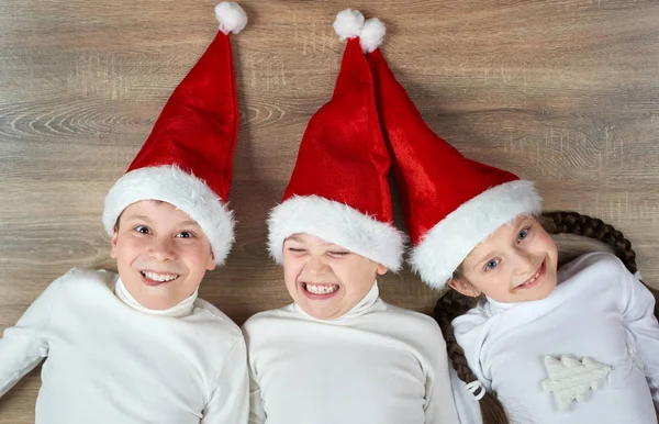 Tres niños en los sombreros de Santa que mienten en el fondo de madera, teniendo emociones divertidas y felices, concepto de vacaciones de invierno —  Fotos de Stock