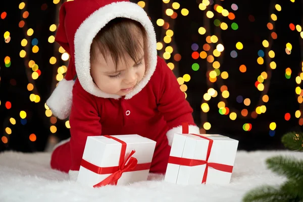 Bebé desempaquetar cajas de regalo con decoración de Navidad, vestido como Santa Claus, luces de arco sobre fondo oscuro, concepto de vacaciones de invierno — Foto de Stock