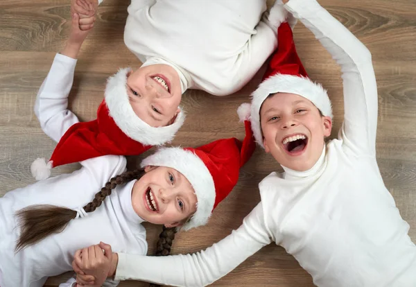Três crianças em chapéus de Papai Noel deitado em fundo de madeira, se divertindo e emoções felizes, conceito de férias de inverno — Fotografia de Stock