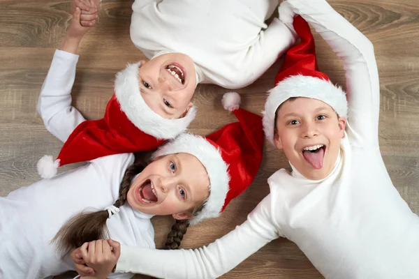 Tres niños en los sombreros de Santa que mienten en el fondo de madera, teniendo emociones divertidas y felices, concepto de vacaciones de invierno — Foto de Stock