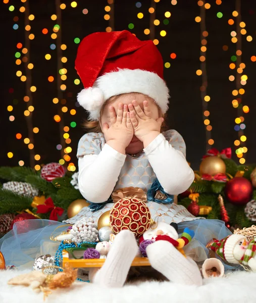 child girl hide face in christmas decoration, happy emotions, winter holiday concept, dark background with illumination and boke lights