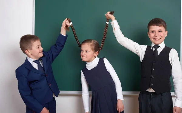 Kinder spielen und Spaß haben, Jungen ziehen die Zöpfe der Mädchen, in der Nähe leere Schultafel Hintergrund, im klassischen schwarzen Anzug gekleidet, Gruppenschüler, pädagogisches Konzept — Stockfoto