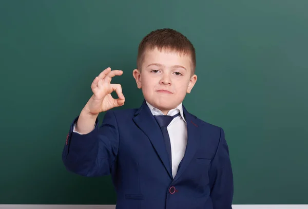School boy show ok sign, portrait near green blank chalkboard background, dressed in classic black suit, one pupil, education concept — Stock Photo, Image