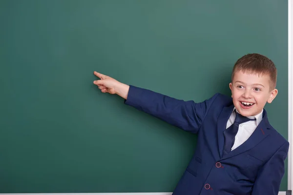 Schuljunge zeigt mit dem Finger in die Nähe eines leeren Tafelhintergrundes, gekleidet in einen klassischen schwarzen Anzug, Gruppenschüler, pädagogisches Konzept — Stockfoto