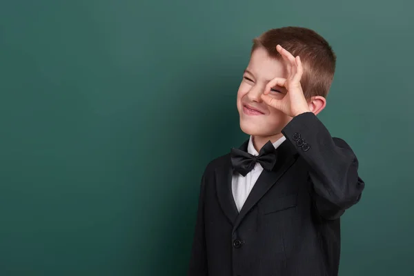 Escuela chico mostrar ok signo, retrato cerca verde en blanco pizarra fondo, vestido con traje negro clásico, un alumno, concepto de educación — Foto de Stock