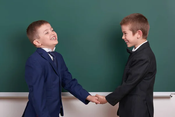 Dwóch przyjaciół uścisk dłoni, chłopiec szkoły podstawowej w pobliżu pusta tablica tło, ubrany w klasyczny czarny garnitur, grupa uczniów, koncepcja edukacji — Zdjęcie stockowe
